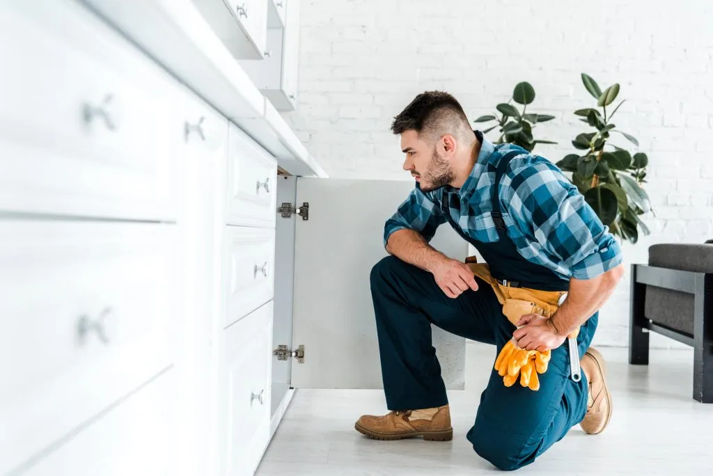 Selective focus of handsome handyman with tool belt sitting near a kitchen cabinet